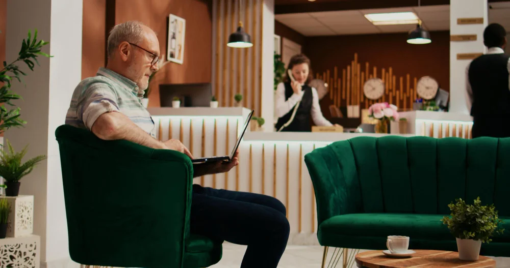 An older man reading a book in a cozy room with green chairs.